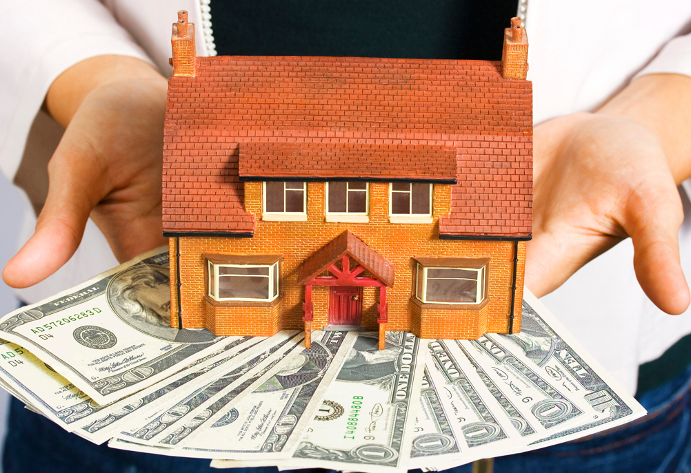 A person holding a model house resting on a stack of US dollar bills.